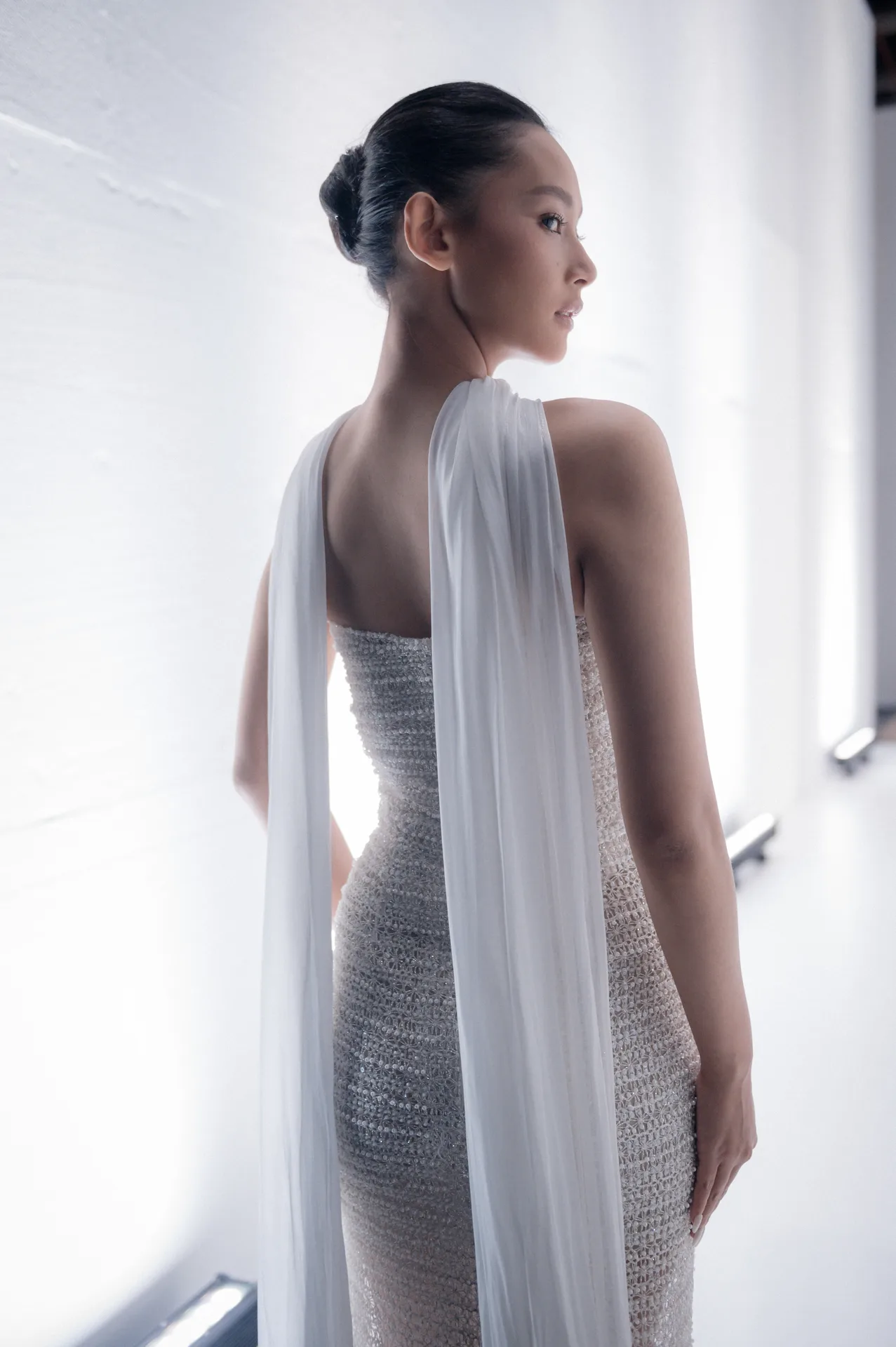 Model looking over shoulder in hallway wearing textured silver strapless gown with white shoulder drapes