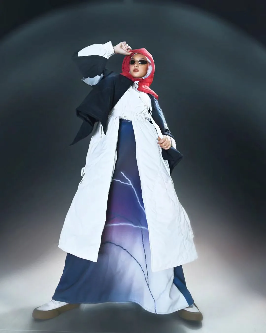 Runway model wearing red headpiece and oversized white-and-blue graphic long coat under spotlight