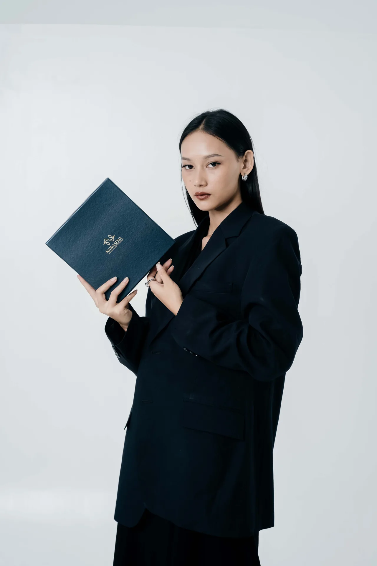 Studio lookbook portrait of female model in oversized black blazer holding navy blue diploma folder