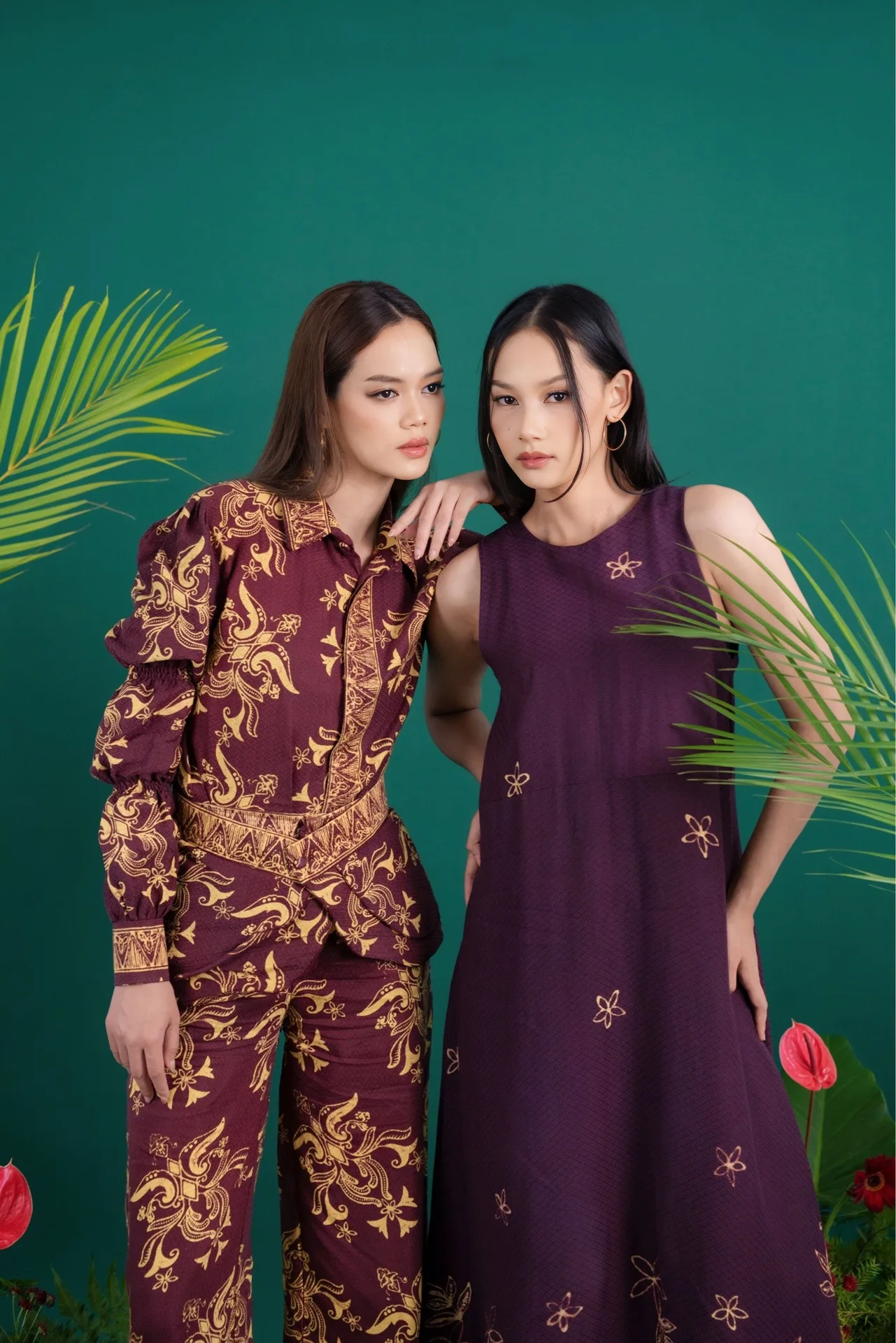 Two female models posing in maroon and purple batik patterned outfits against green studio backdrop