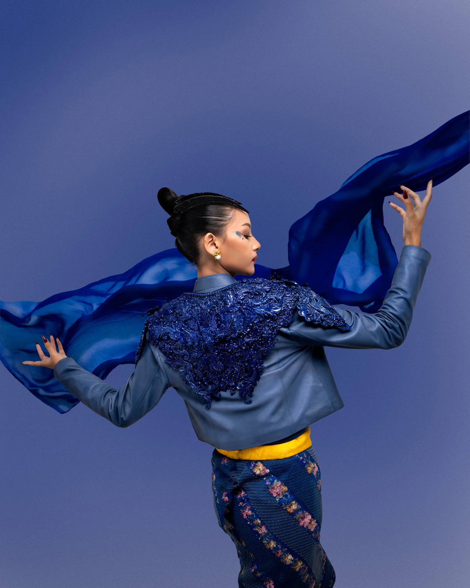 Model in cobalt blue jacket holding aloft fluttering sheer blue silk fabric against periwinkle backdrop